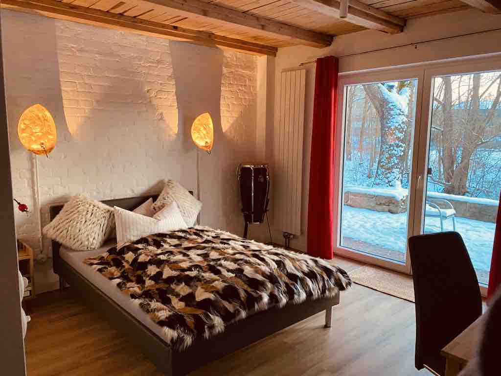 An inviting sleeping area features a spacious bed adorned with a textured throw blanket. Natural light streams through large sliding doors, revealing a snowy outdoor setting. Two contemporary lamps provide soft lighting, while a pair of drums adds an artistic touch to the room.