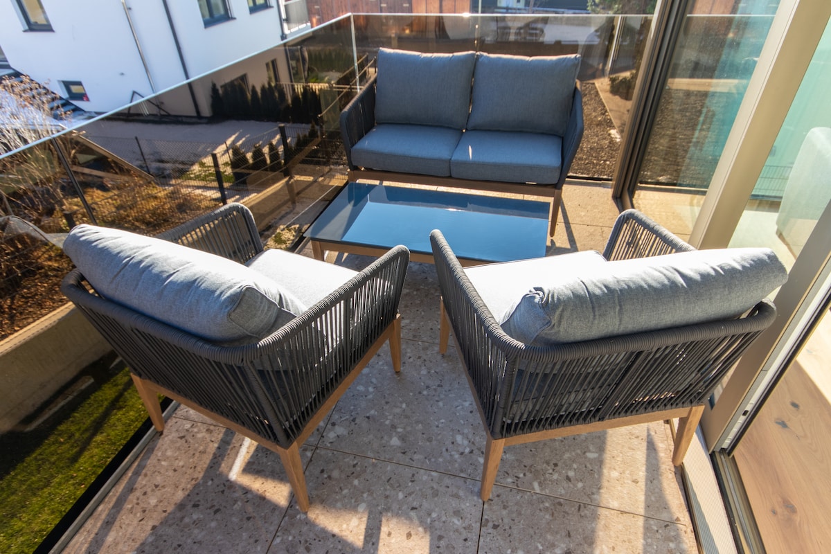 An outdoor seating area features two lounge chairs and a sofa, all with soft, gray cushions. A glass table sits in the center on a stone patio, surrounded by green landscaping, creating a comfortable space for relaxation.