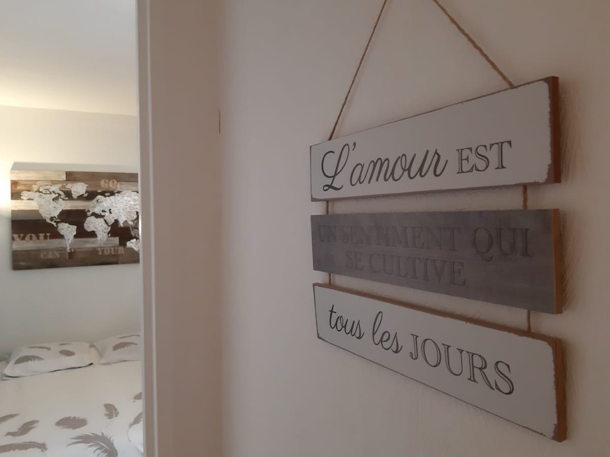 A wall sign with the French phrase "L'amour est un sentiment qui se cultive tous les jours" is displayed near a bedroom entrance. The background features a textured artwork of a world map above a cozy bed, creating an inviting entryway to the sleeping area.