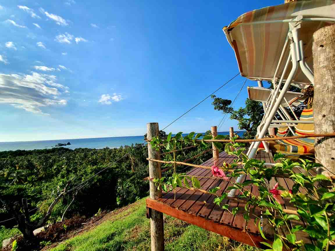 A wooden deck features comfortable chairs positioned to overlook a lush landscape that extends to the ocean. Colorful flowers adorn the railing, and the sky is bright with a few clouds, enhancing the tranquil outdoor setting.