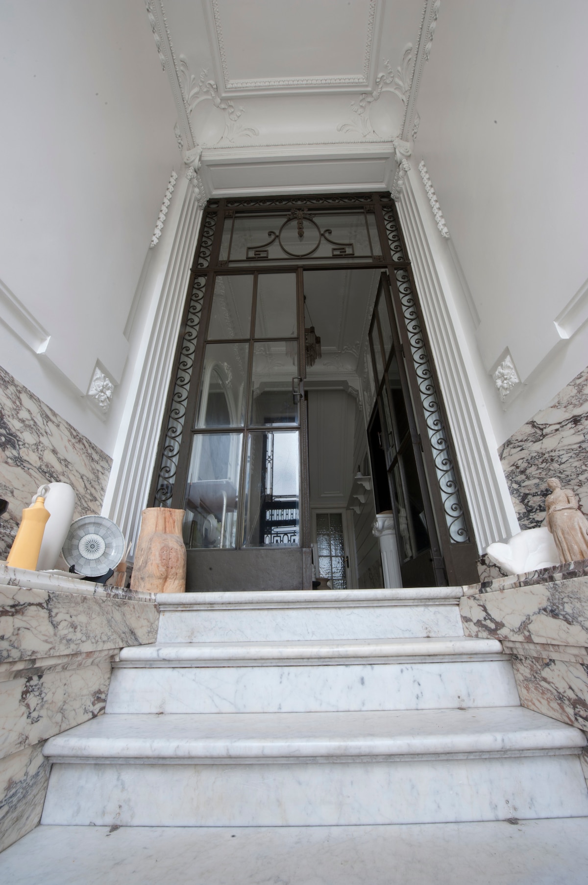 A grand entrance showcases a stately marble staircase leading up to double glass doors. The ornate door design incorporates geometric patterns, while decorative elements are present beside the steps, including vases and sculptures. Natural light filters through the entrance, enhancing the architectural details.