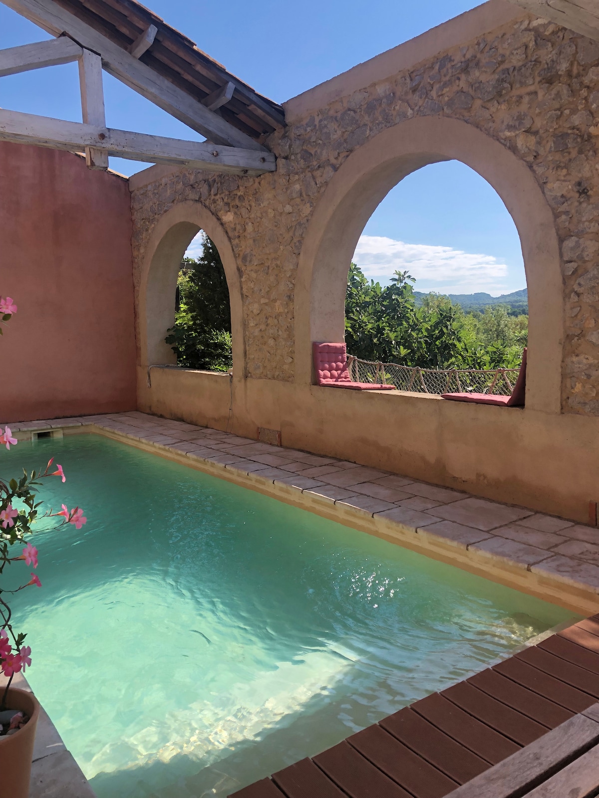 A serene swimming pool is framed by arched stone walls, with sunlight reflecting on the water's surface. Potted flowers add a touch of color, and lush greenery is visible beyond the pool, providing a natural backdrop.