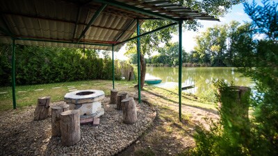 A covered fire pit area is surrounded by rustic log seating, providing a space for gatherings. The landscape features lush greenery and a calm waterbody visible just beyond. A boat appears moored nearby, enhancing the natural setting.