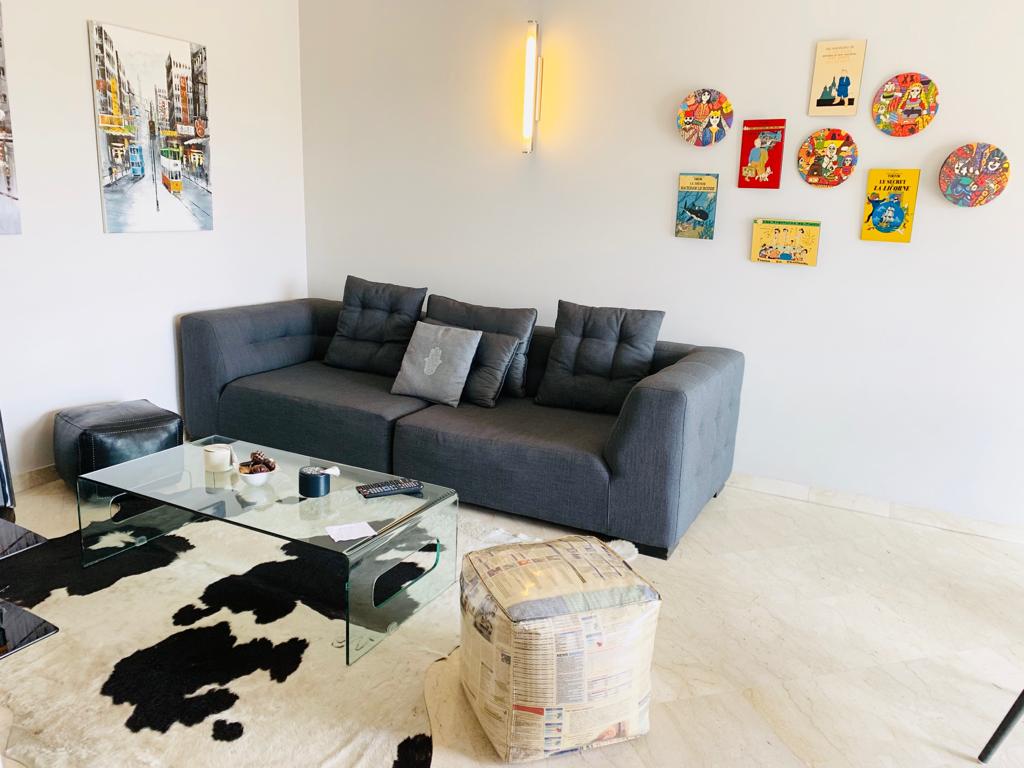 A modern living area features a dark gray sofa with decorative cushions, complemented by a glass coffee table. Vibrant wall art, including colorful plates, adorns the wall, while a plush black and white rug adds texture to the floor.