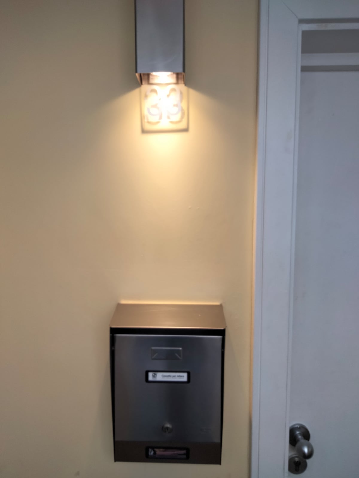 A wall-mounted mailbox is positioned beneath a contemporary light fixture. The mailbox features a sleek metal design and is labeled with a clear address number. Soft light illuminates the wall, enhancing visibility in the entryway.