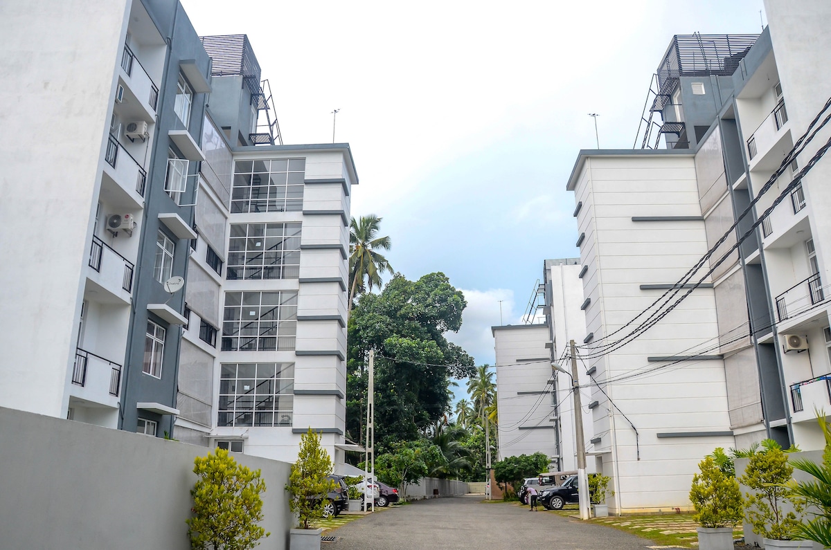 A view of the two adjoining apartment buildings within the complex, showcasing a well-maintained pathway lined with greenery. The structures are modern with large windows and balconies, offering a sense of spaciousness and accessibility.