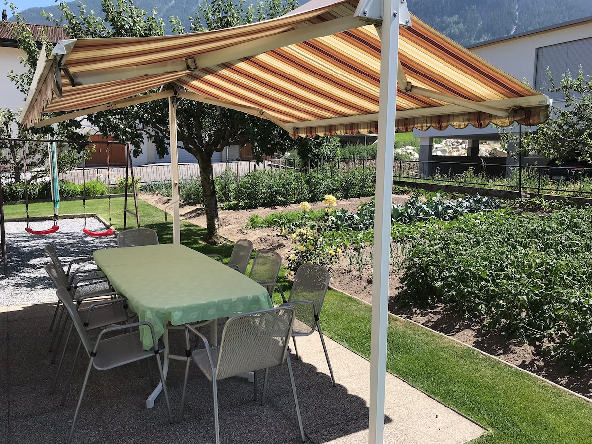 An outdoor dining area features a long table covered with a light green cloth, surrounded by several metal chairs. An awning provides shade over the setting, and a grassy area is visible alongside a garden with various plants. Red swings are seen in the background.