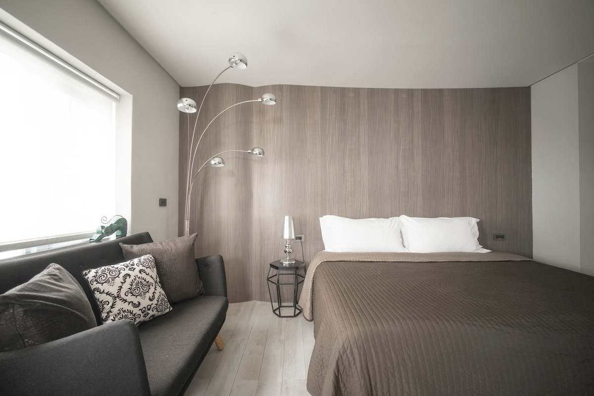 A modern sleeping area features a spacious bed with neutral bedding against a softly curved wooden wall. A sleek sofa with decorative cushions complements the space. A stylish floor lamp stands nearby, providing gentle illumination. Natural light enters through a window covered by sheer blinds.