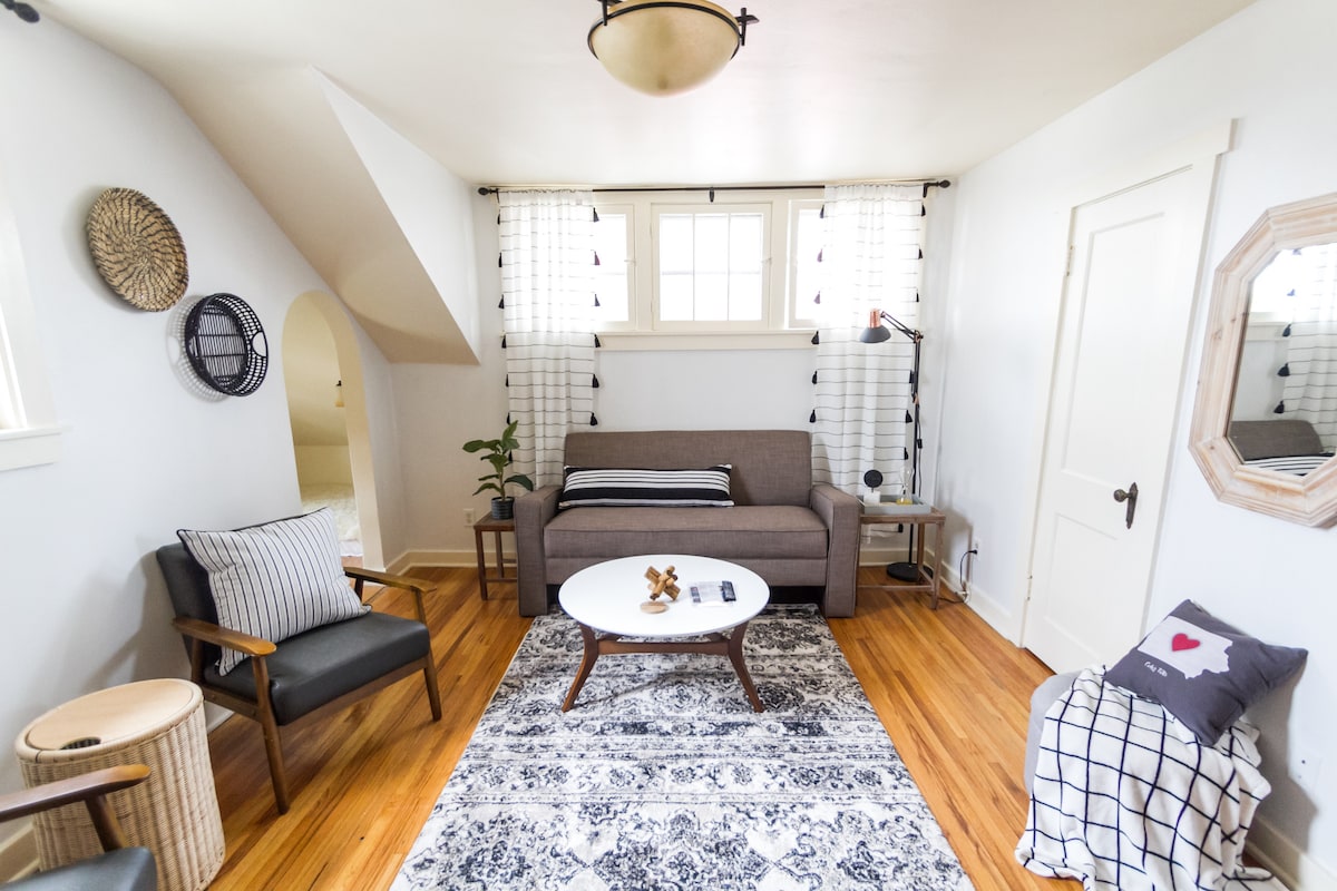 A living area features hardwood flooring and warm lighting. A comfortable sofa is positioned beneath natural light from large windows, surrounded by decorative curtains. A round coffee table rests on a patterned area rug, while a plant and various decorative items enhance the space.