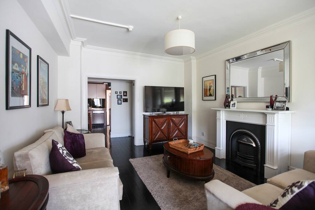 Magnificent high-ceiling Art Deco living room (including artwork to match) with views of Sydney Harbour, a faux-fireplace, huge comfy couches, antique writing desk, and a 60-inch high-definition Samsung TV (connected with the new Apple TV - perfect for a movie night in with your feet up on the extra-wide ottoman!)