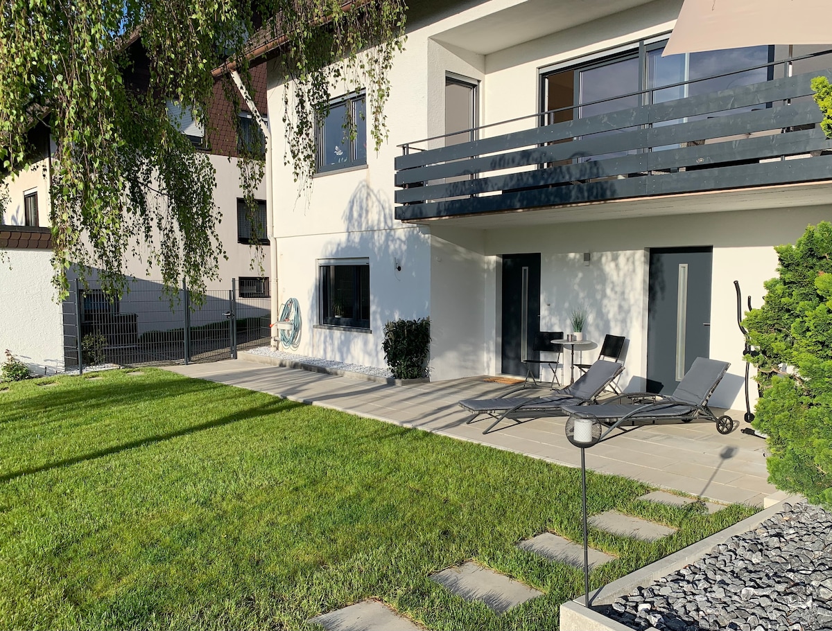 A patio area is shown with a well-maintained lawn. Comfortable lounge chairs are positioned for relaxation, alongside a small table. A section of the building with a balcony is visible, framed by greenery and natural light.