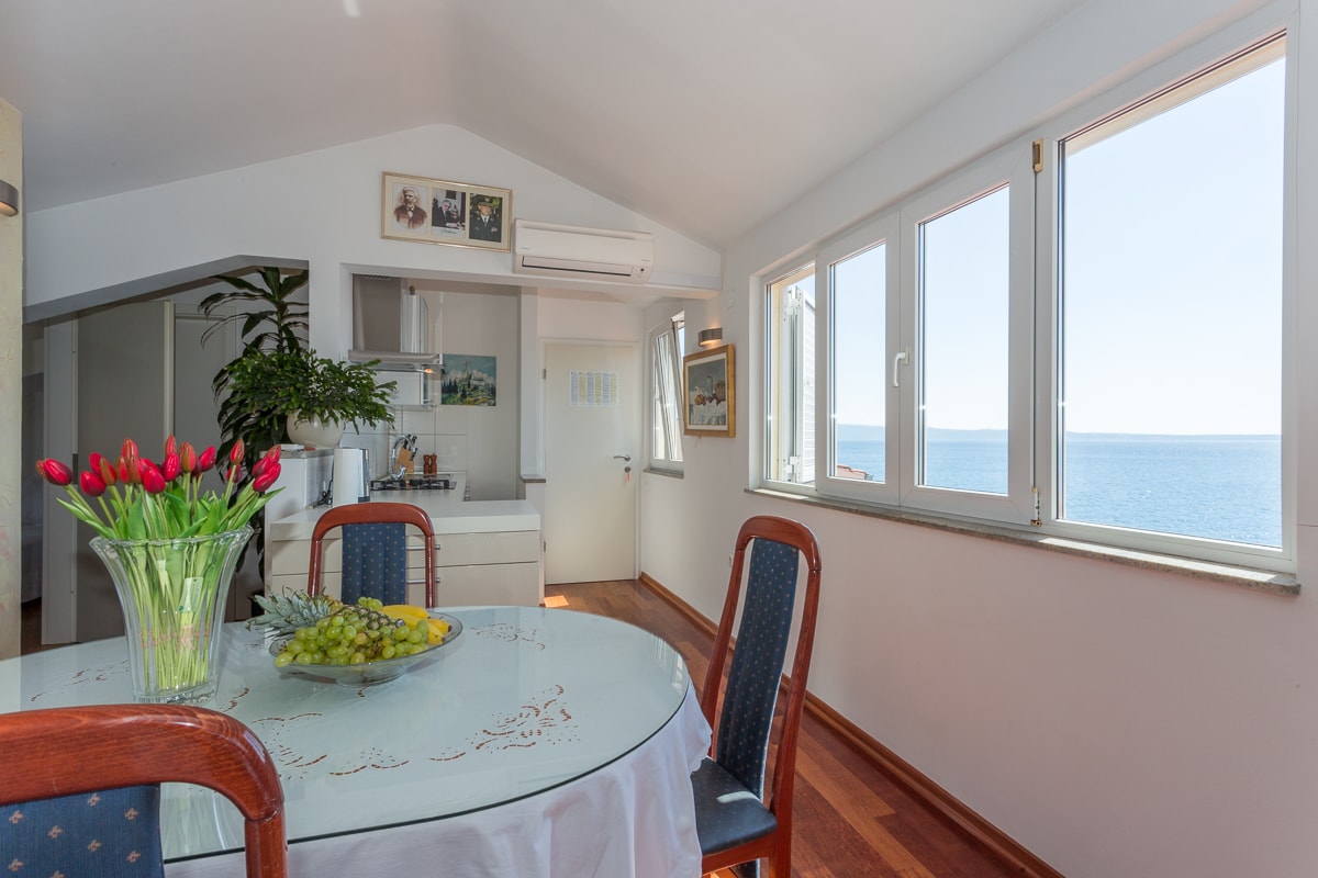 A bright dining area features a round table set for four, adorned with a floral tablecloth and a vase of tulips. Large windows provide a view of the sea, filling the space with natural light, while a kitchen area is visible in the background.