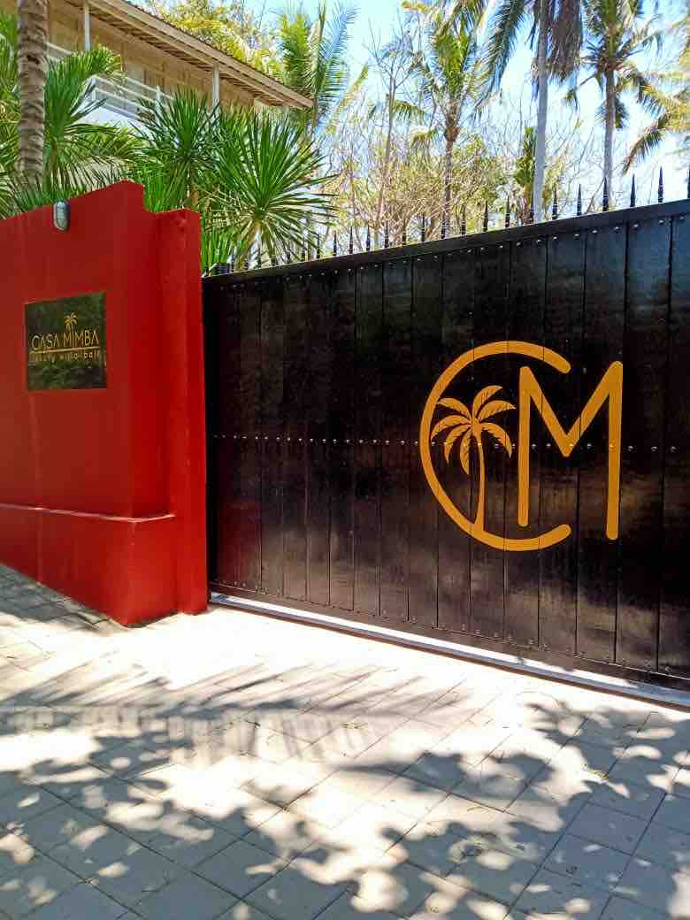 A striking entrance gate showcases a bold logo featuring palm trees. The gate is framed by vibrant red walls and tropical greenery, providing a welcoming ambiance to the villa. Sunlight filters through the nearby palm leaves, creating dappled shadows along the pathway.