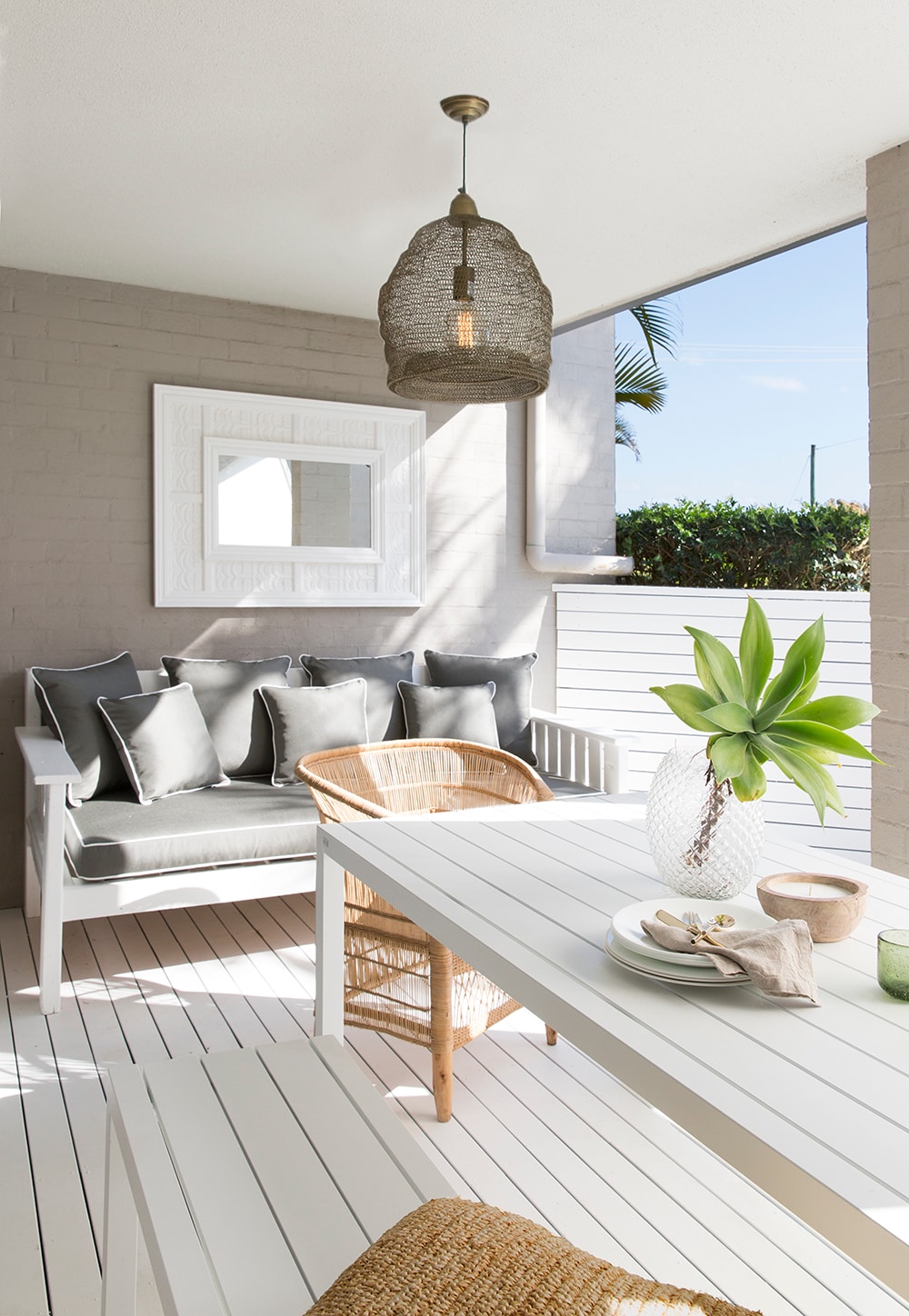 A stylish outdoor seating area features a light-hued table and two chairs, including a rattan one. A cushioned bench with decorative pillows offers comfortable seating. Natural light illuminates the space, while a pendant light hangs above, and potted greenery is visible in the background.