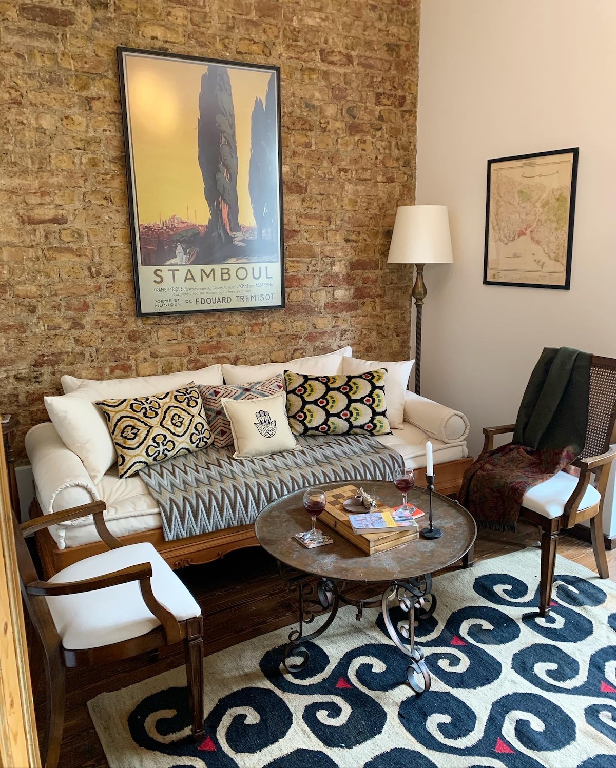 A cozy corner of the living area features an exposed brick wall adorned with a vintage poster. A white sofa with decorative cushions is paired with two accent chairs. A small round table showcases a candle and books, inviting relaxation.