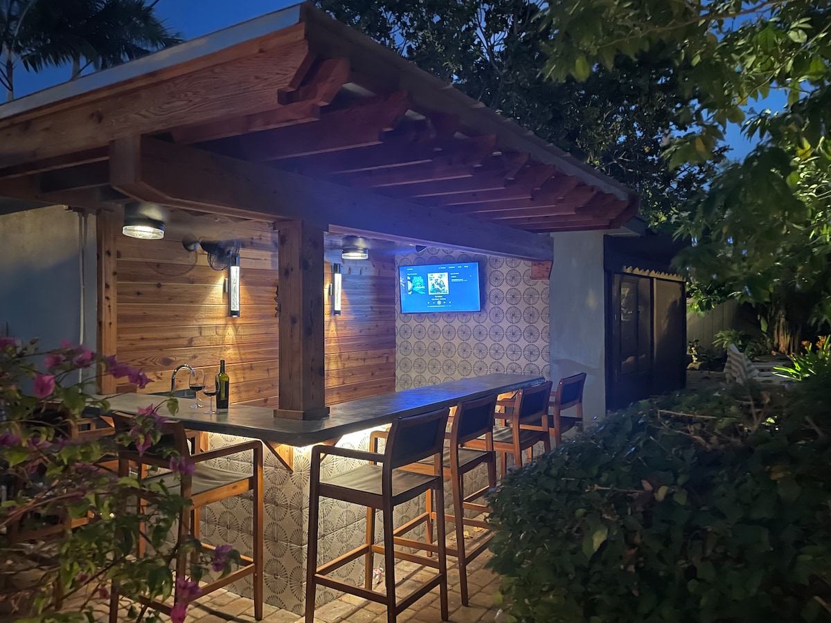 An elevated outdoor bar is featured under a wooden structure, surrounded by lush greenery. Bar stools are arranged along the countertop, and a flat screen TV is mounted on the wall, enhancing the space for gatherings. Soft lighting creates a cozy atmosphere in the evening.