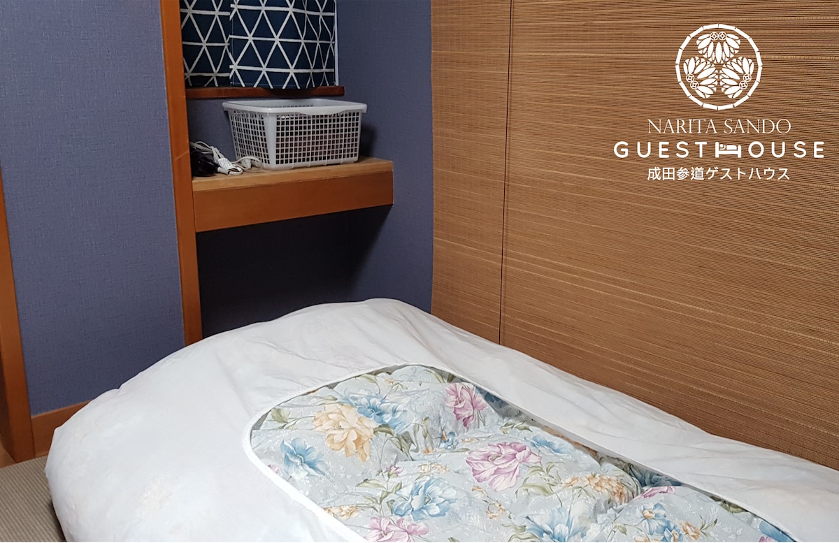 A cozy Japanese-style sleeping area is presented with a spacious futon placed on a tatami mat. Nearby, a low wooden shelf holds a small basket, offering storage options. The walls are adorned in soft blue and warm wood tones, creating a serene environment.