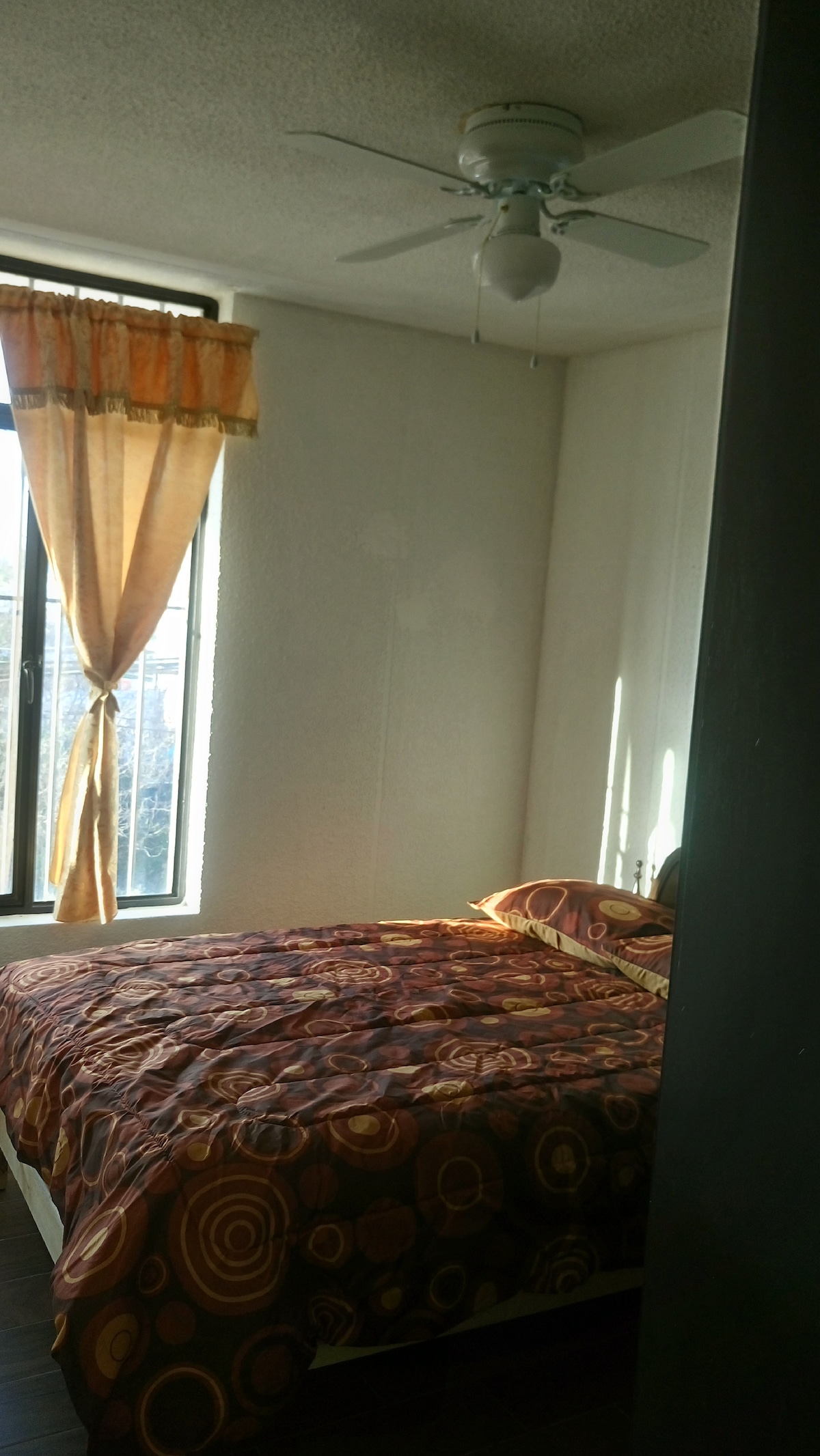 A well-lit bedroom features a spacious bed with an ornamental cover in warm tones. A soft, sheer curtain frames the window, allowing natural light to fill the room. A ceiling fan is positioned overhead, providing ventilation.