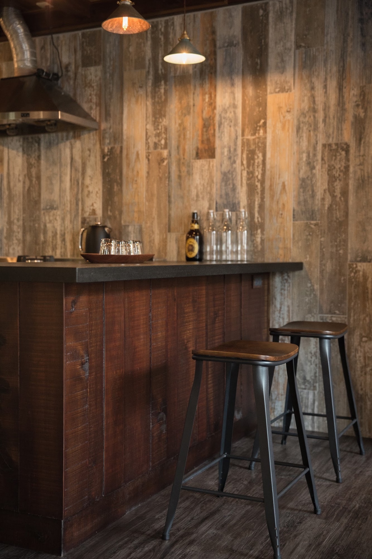A rustic bar area features a dark wood countertop backed by a textured wood accent wall. Two metal stools with wooden seats are positioned at the bar. Glass bottles and a ceramic container are displayed on the countertop, complemented by warm lighting from pendant fixtures.
