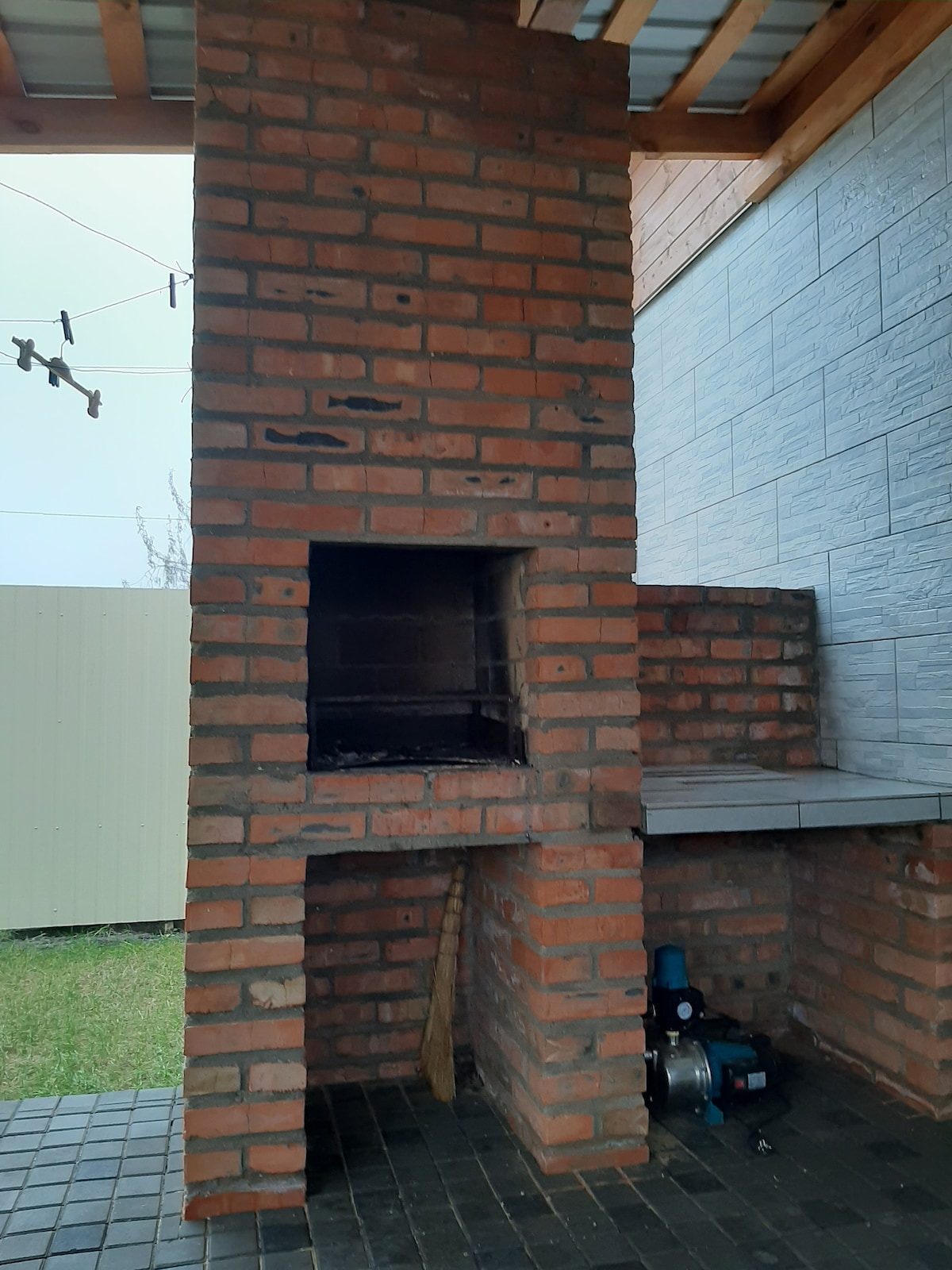 A brick barbecue grill is shown against a wall, featuring an open cooking area and a smoke vent. A stone surface extends alongside the grill, while a green lawn is visible in the background. Clothes are hung on a line, indicating outdoor use.