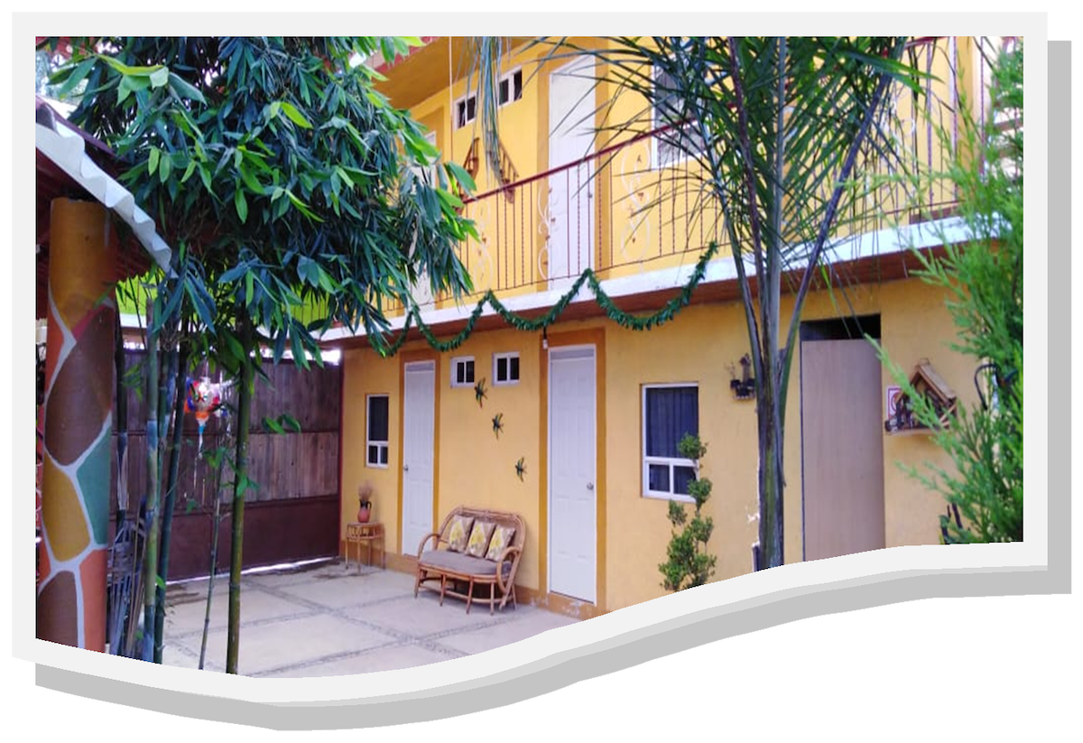 The image features a two-story building with a yellow exterior, surrounded by greenery. A small seating area with a wooden couch is visible next to a white entrance door. Decorative garlands add a festive touch to the balcony area.