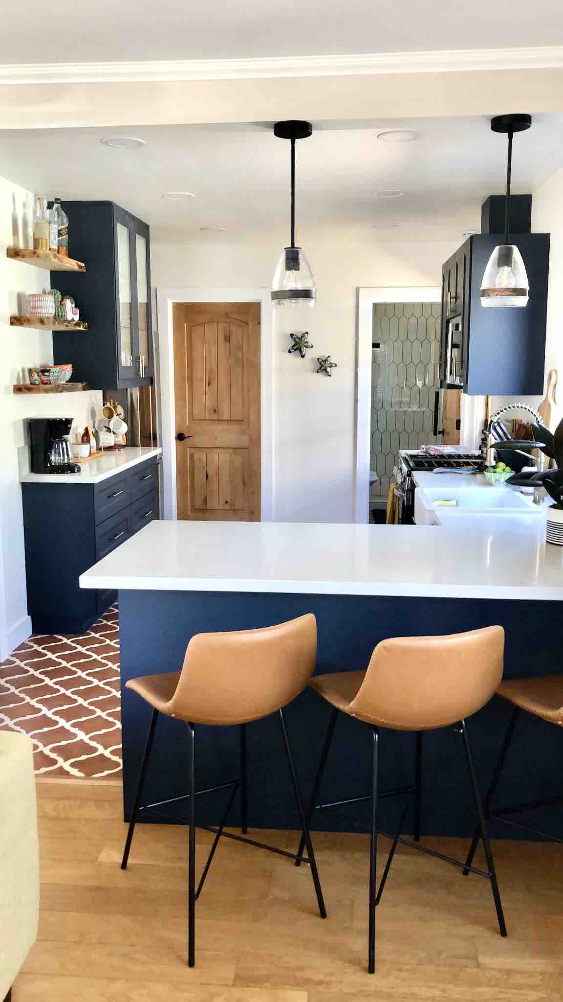 A modern kitchen setup is featured, showcasing a dark blue cabinetry, white countertops, and sleek pendant lighting. Two brown bar stools are positioned at the kitchen island, with open shelving displaying kitchen essentials. A wooden door leads to another area of the home in the background.