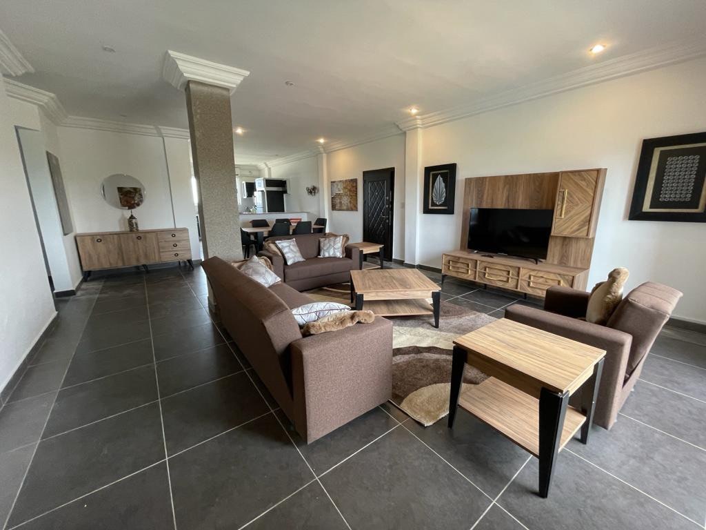 A modern living area is presented with comfortable brown sofas and accent chairs arranged around wooden coffee tables. The floor features dark tiles, and a large wall-mounted television is visible. Art pieces adorn the walls, adding character to the space.