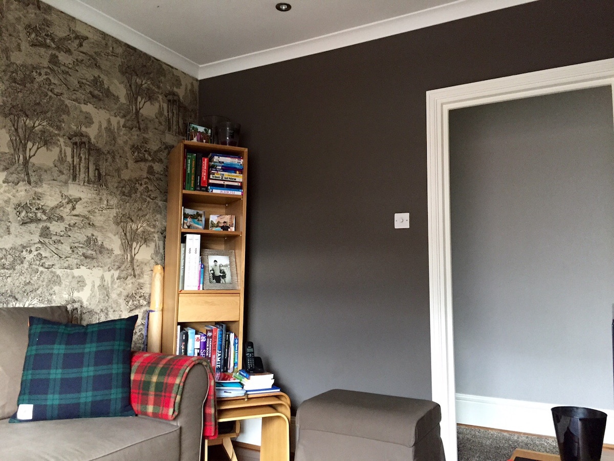 The living room features a neutral color scheme, with a large comfortable sofa adorned with a plaid throw. A wooden bookshelf filled with books stands against the wall, complemented by a soft ottoman. Natural light enters through the adjacent doorway, enhancing the inviting space.