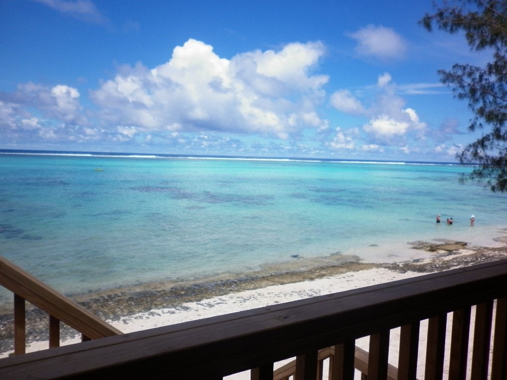 A view of the tranquil sea is presented, showcasing vibrant blue waters and a sandy shoreline. A wooden railing frames the perspective, while clouds drift softly in the sky. A few individuals are observed wading near the water, emphasizing the beach's inviting atmosphere.