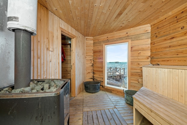 A rustic sauna interior features natural wood walls and a wooden floor. A stone stove stands prominently, with a stack of stones atop. The large window offers a view of the waterfront, complementing the inviting atmosphere created by warm wood tones.