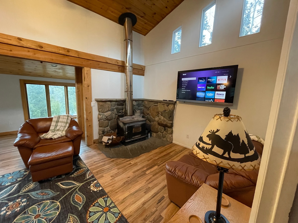A spacious living area features warm wood tones with a stone woodstove as the focal point. Comfortable leather seating is arranged for relaxation. A large television is mounted on the wall, and large windows provide natural light and views of the surrounding nature.