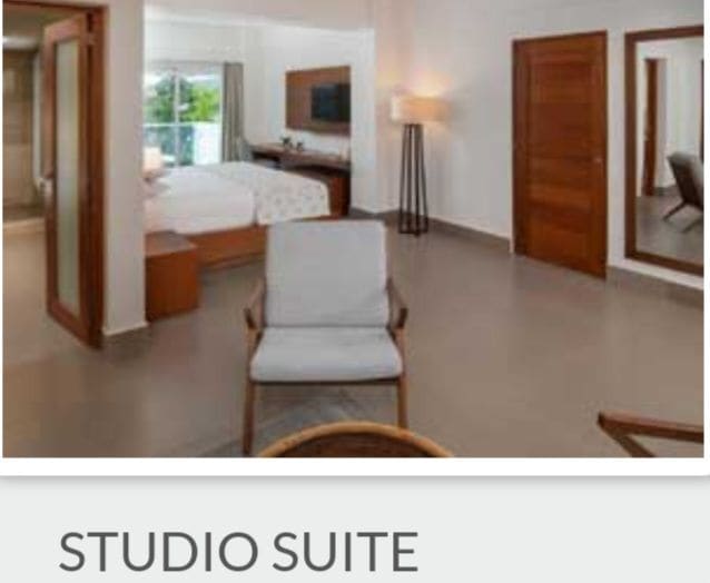 A spacious junior studio suite features a king bed with a cozy bedspread, positioned next to large windows that allow natural light. A comfortable chair is placed in the foreground, while a well-appointed bathroom is visible through an open door.