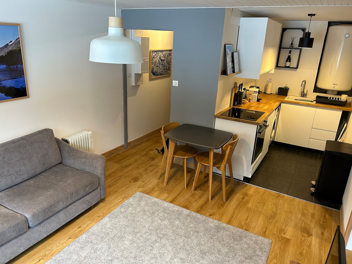 A modern living and dining area features a gray sofa and a round black dining table accompanied by two wooden chairs. A well-equipped kitchen with sleek cabinets and appliances is visible in the background, complemented by wooden flooring and a light fixture hanging from the ceiling.