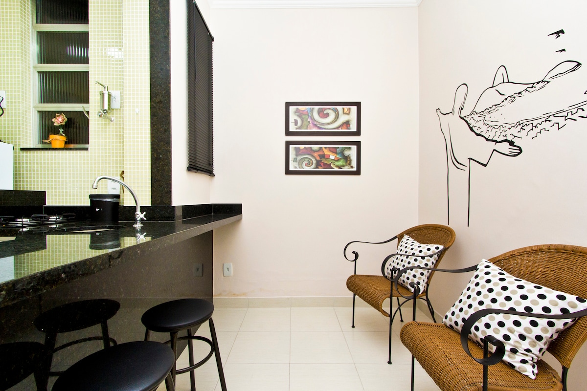 An open kitchen area is visible, featuring dark granite countertops and sleek black bar stools. Two wicker chairs with polka dot cushions are positioned nearby, creating a casual seating space. The wall is adorned with black and white artwork, adding character to the room.