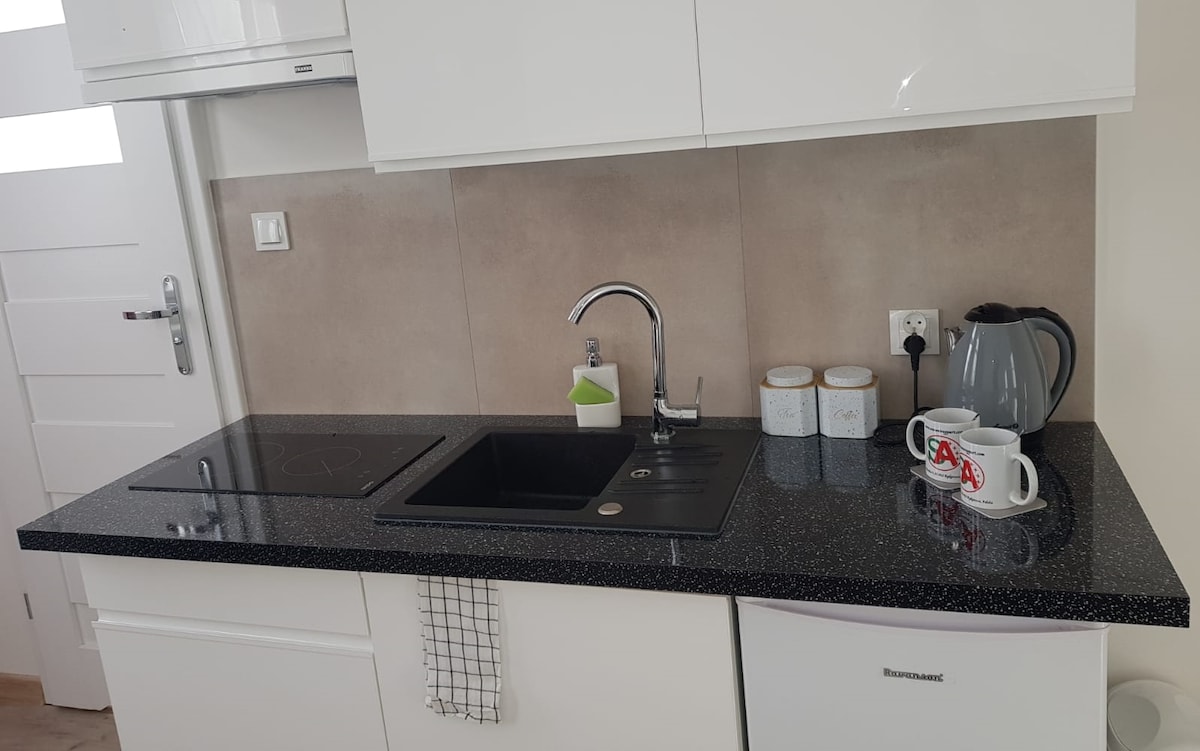 The kitchen area features a sleek black countertop with a stove and a sink. White cabinetry complement the modern design. A kettle and several mugs are displayed alongside three storage jars, with a compact refrigerator positioned beneath the counter.