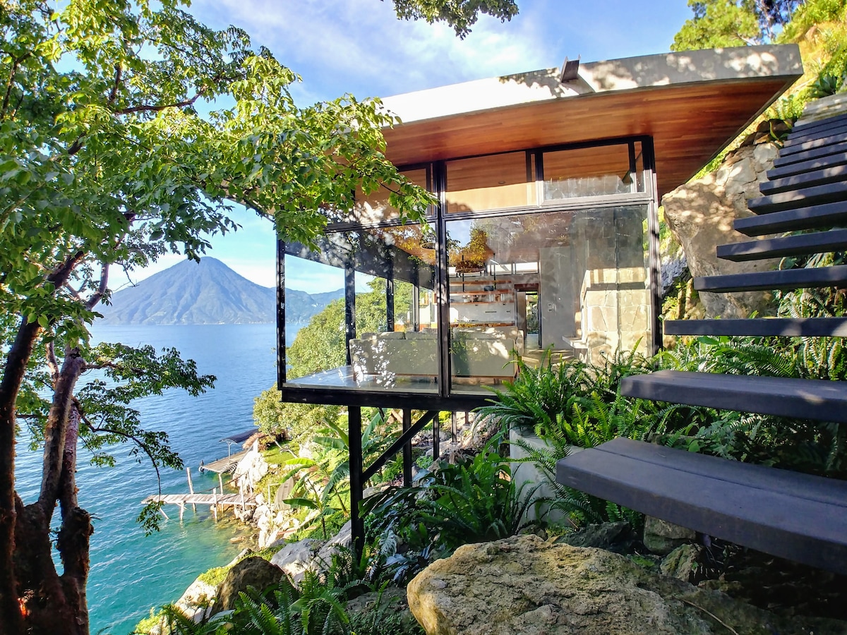 The modern minimalist home features floor-to-ceiling glass walls that enhance views of Lake Atitlan and the surrounding greenery. Steps leading up to the entrance are flanked by lush plants, while the shoreline and dock can be seen in the background.