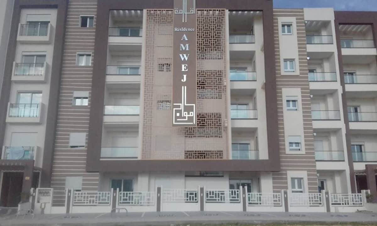 The exterior view of a modern residential building features a multi-level façade with a contemporary design. The structure includes several balconies and large windows, framed by a subtle color palette. The entrance is marked by a decorative sign bearing the name of the residence.