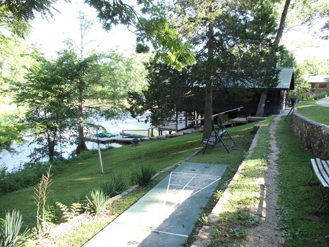 A lush green lawn leads to a serene lakefront, where a wooden dock is visible. Canoes are stationed near the water, and a pathway lined with benches encourages relaxation. Tall trees provide shade, creating a peaceful outdoor environment.