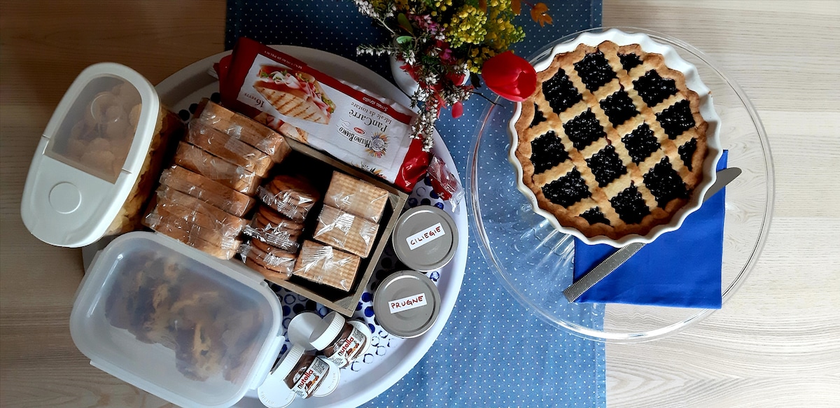 A circular tray displays a variety of breakfast items, including a pie topped with dark fruit, an assortment of packaged cookies, and jars of spread. A small vase with flowers is positioned beside the tray, adding a touch of colour to the presentation.