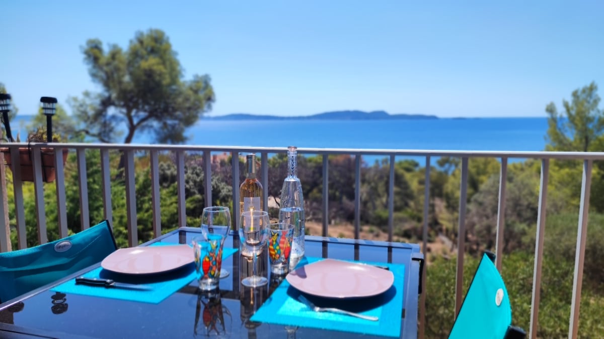 A terrace dining setup is presented, featuring a glass table adorned with plates and glasses. The backdrop showcases a stunning view of the sea and distant islands, framed by lush greenery. Blue table accents complement the serene coastal atmosphere.
