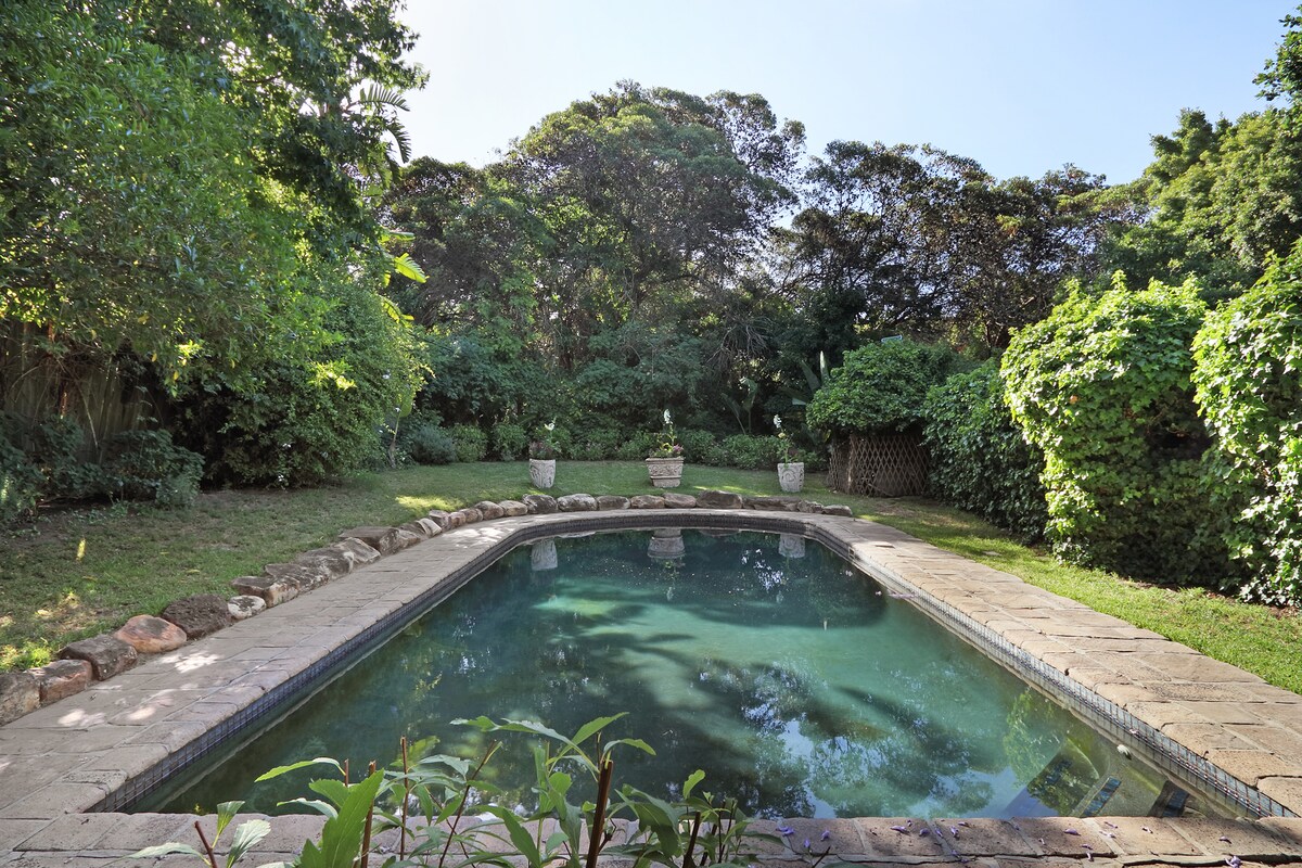 A shared pool is surrounded by lush greenery and shrubs, offering a serene outdoor space. Stone tiles frame the pool, while potted plants are placed strategically around the edge, contributing to a sense of tranquility.