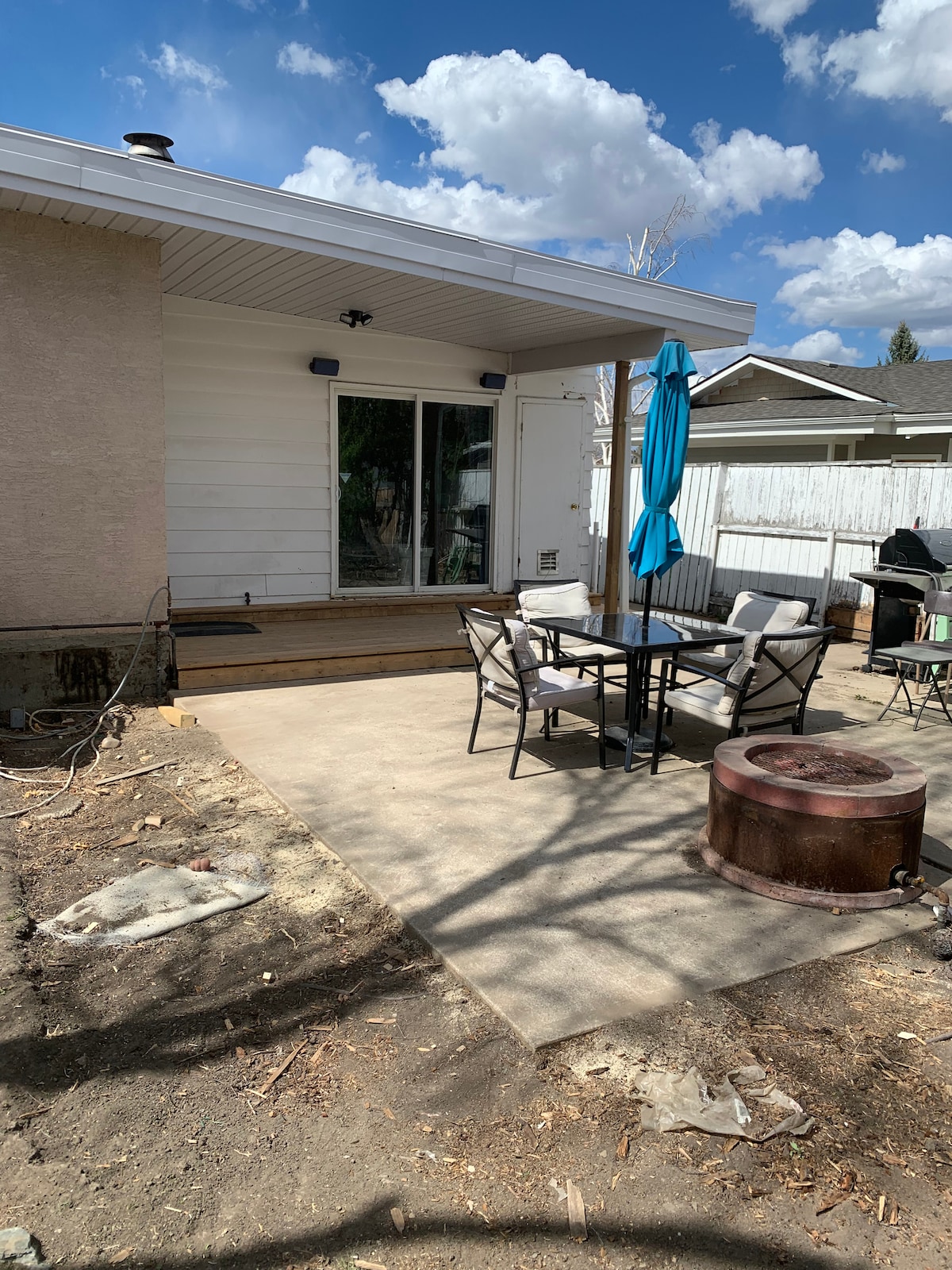 An outdoor patio features a round table with four chairs under a blue umbrella. A small fire pit is visible on a stone surface, with a concrete area beside it. The space appears to be well-lit, surrounded by greenery and a fenced yard.