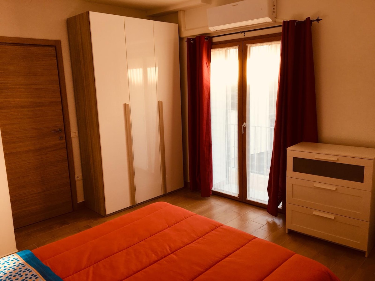 A bright bedroom featuring a spacious bed covered in an orange bedspread. A large wardrobe with sliding doors is positioned next to a chest of drawers. Natural light streams through the glass door leading to a small balcony, framed by red curtains.