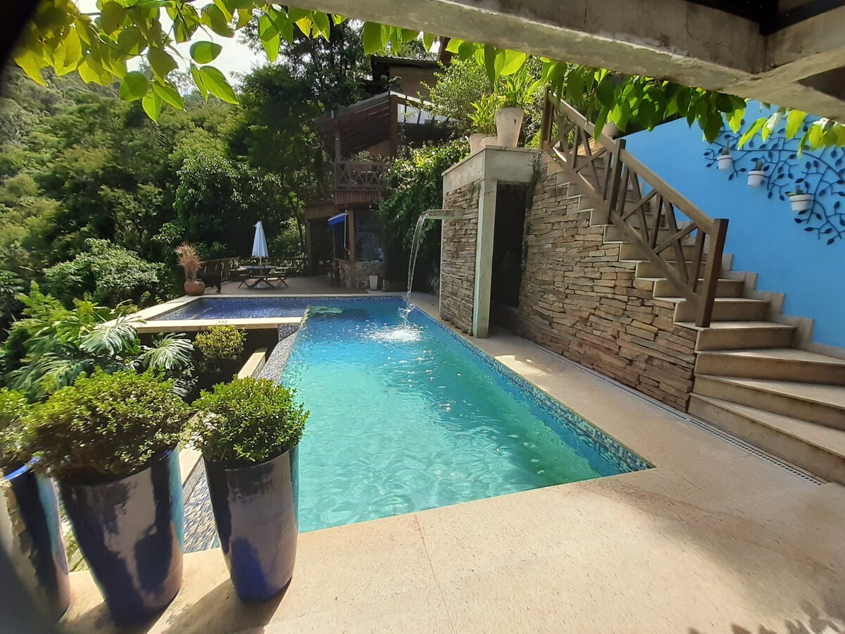 A serene outdoor area features a sparkling pool with a gentle waterfall. Surrounded by lush greenery, potted plants, and a view of a staircase leading to a decked area, the tranquil space invites relaxation and leisure.