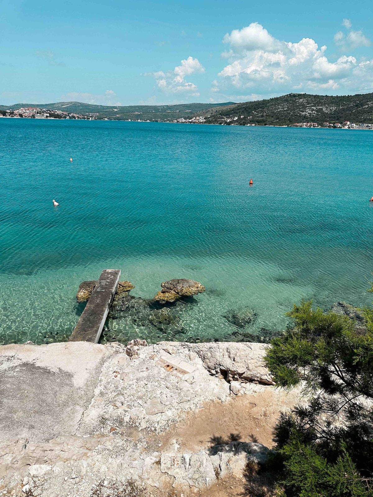 The image captures a serene coastal view with clear turquoise waters gently lapping against the rocky shore. A wooden jetty extends out into the water, surrounded by smooth stones. Lush greenery partially frames the scene, while distant hills can be seen under a clear blue sky.