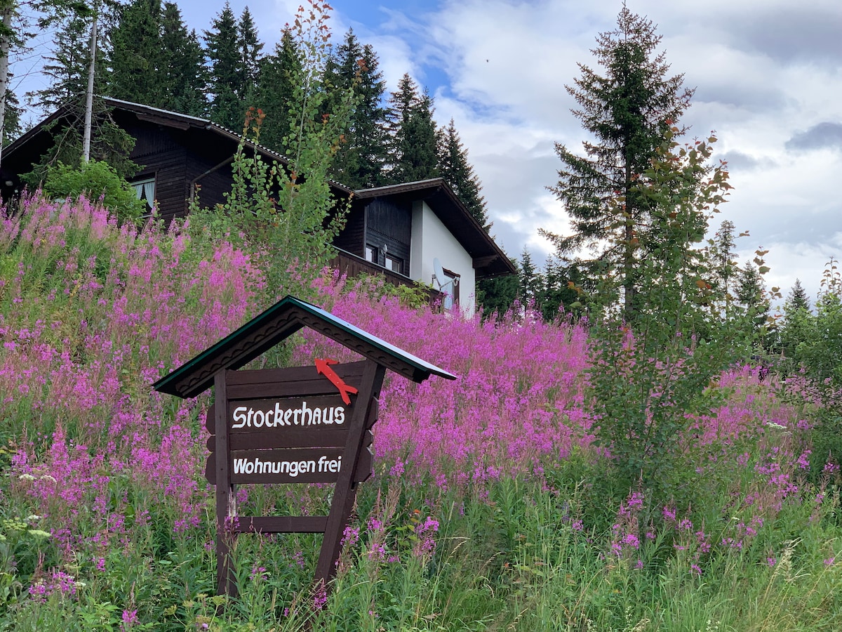 The exterior of Stockerhaus is visible, framed by vibrant purple wildflowers. A wooden sign displays the name 'Stockerhaus' and the text 'Wohnungen frei.' The two-story building is surrounded by lush greenery and tall trees against a cloudy sky.
