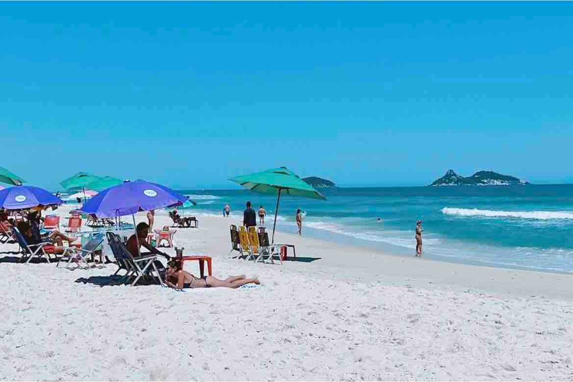 The beach setting features a broad expanse of soft, white sand, dotted with colorful umbrellas providing shade. A mix of beach chairs is arranged under the umbrellas, with some individuals relaxing or walking along the shore. Gentle waves can be seen lapping at the coastline.