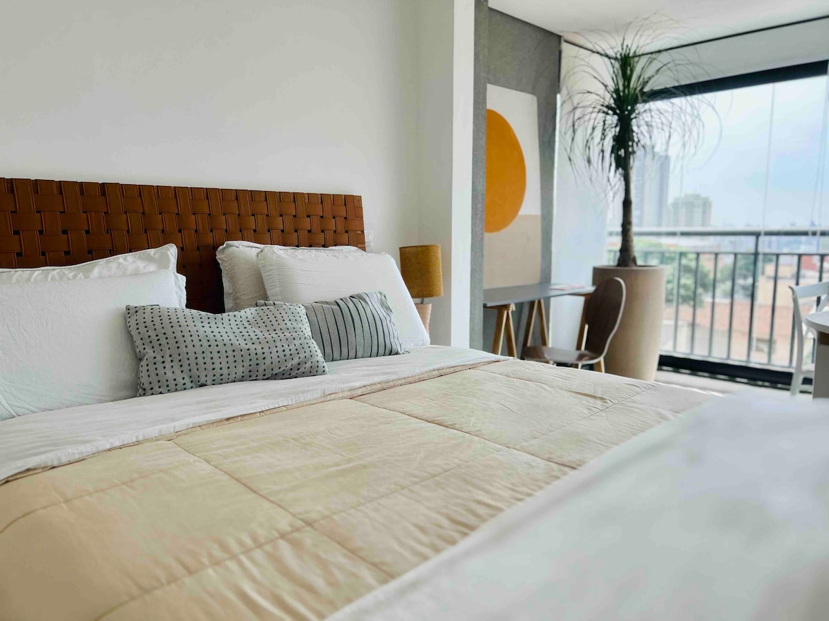 A comfortable sleeping area features a woven headboard and layered bedding. Accent pillows are arranged on the spacious bed. A large window offers a view of the surroundings, while natural light brightens the room. A decorative plant and a modern chair complete the inviting space.