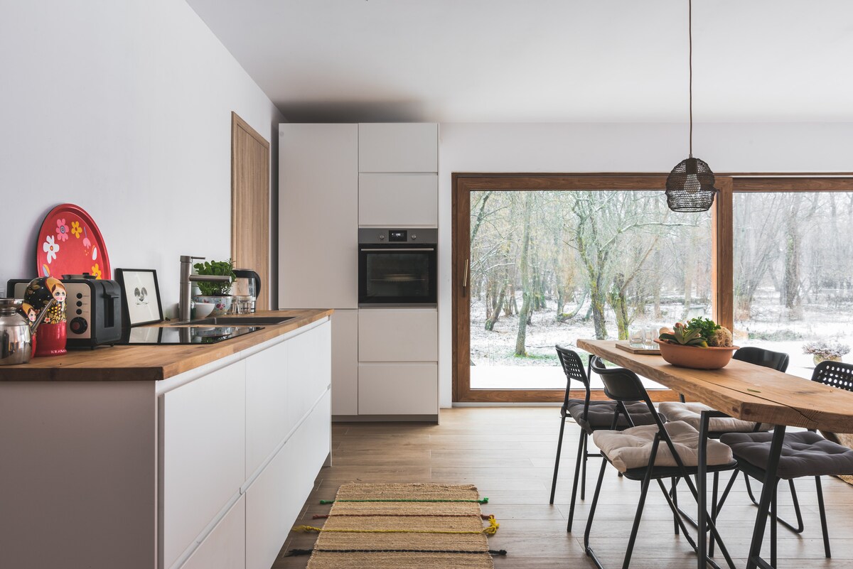A modern kitchen features sleek cabinetry with a wooden countertop. A range oven is integrated into the cabinetry. Light streams through large windows, revealing a natural outdoor setting with trees and snow. A dining table is set with black chairs, and decorative elements are present.