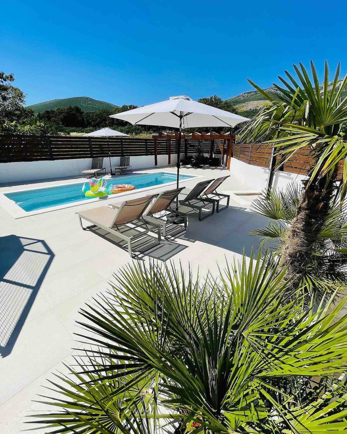 A private pool area features a small heated pool surrounded by lounge chairs and umbrellas. Lush palm trees and greenery frame the space, offering a tropical vibe. The clear blue sky and nearby hills create a serene backdrop for relaxation.